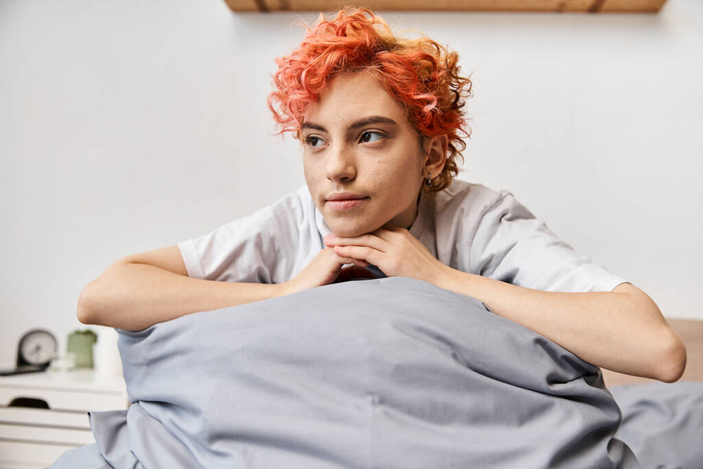 расслабляющий рыжеволосый странный человек в домашней одежде сидит на кровати дома и смотрит вдаль, досуг - Фото, изображение