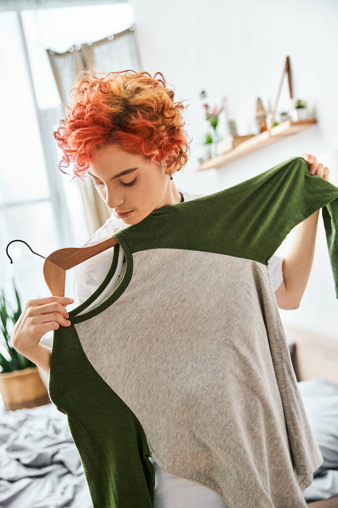 appealing red haired extravagant person picking up stylish clothes near rack at home, leisure time - Photo, Image