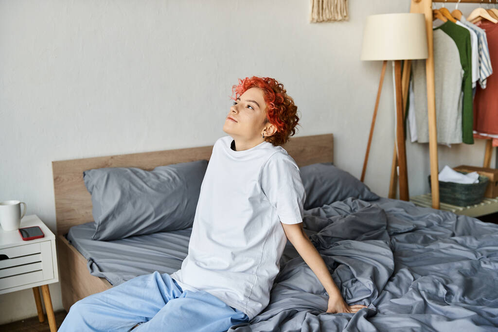 good looking extravagant person in white t shirt with red hair sitting on her bed, leisure time - Photo, Image