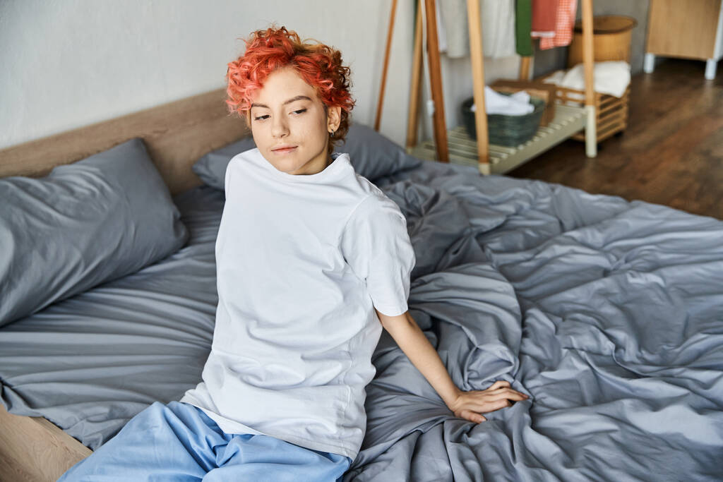 boa aparência pessoa extravagante em t-shirt branca com cabelo vermelho sentado em sua cama, tempo de lazer - Foto, Imagem