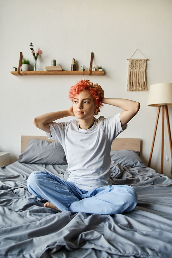 ベッドの上に座って離れて見つめる鮮やかな赤い髪の魅力的なクィア人,余暇時間 - 写真・画像