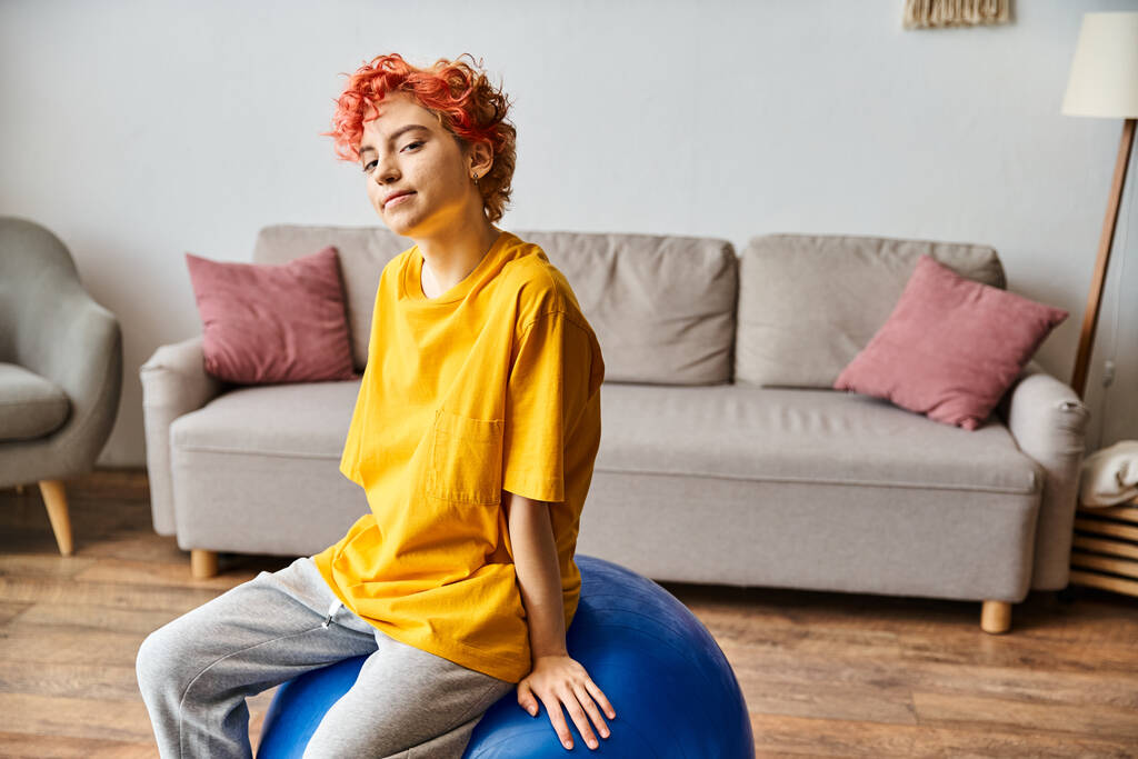 alegre persona queer con el pelo rojo sentado en la pelota de fitness y sonriendo a la cámara mientras está en casa - Foto, Imagen