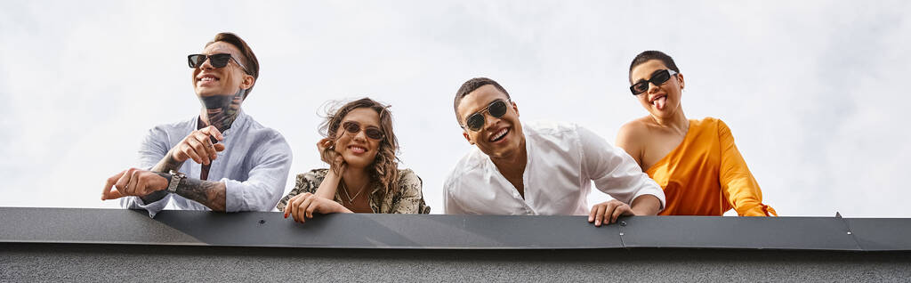 fröhliche multirassische Freunde in urbaner Kleidung mit Sonnenbrille posieren auf dem Dach und lächeln in die Kamera - Foto, Bild