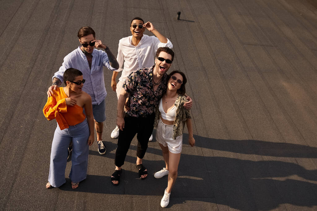 vijf interraciale vrolijke vrienden in levendige kleding poseren op het dak en glimlachen gelukkig op de camera - Foto, afbeelding