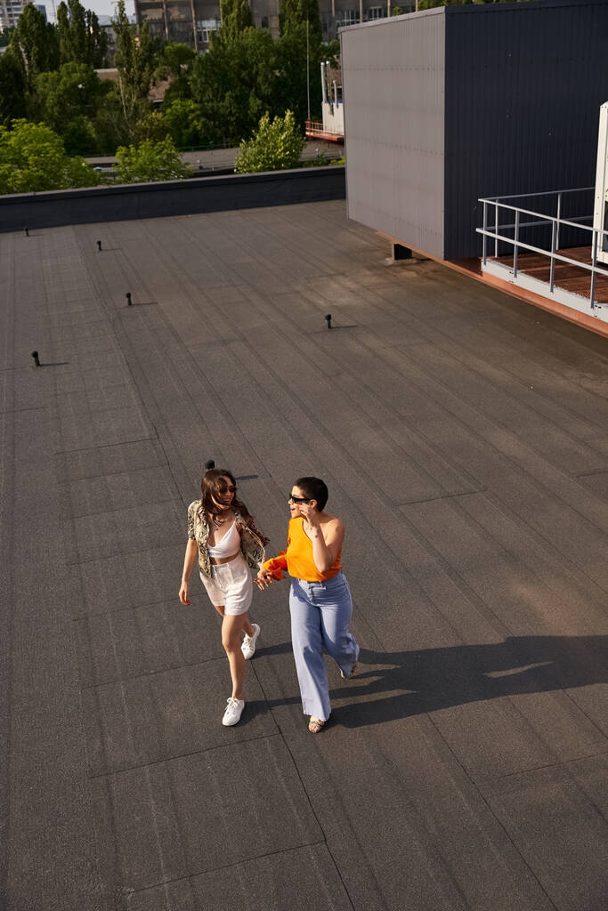 dwie piękne, młode kobiety w luźnych ciuchach w okularach przeciwsłonecznych, pozujące razem na dachu - Zdjęcie, obraz