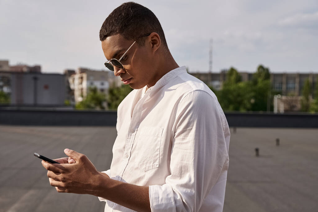 przystojny młody Afroamerykanin człowiek w przypadkowy strój w okulary przeciwsłoneczne patrząc na smartfon - Zdjęcie, obraz
