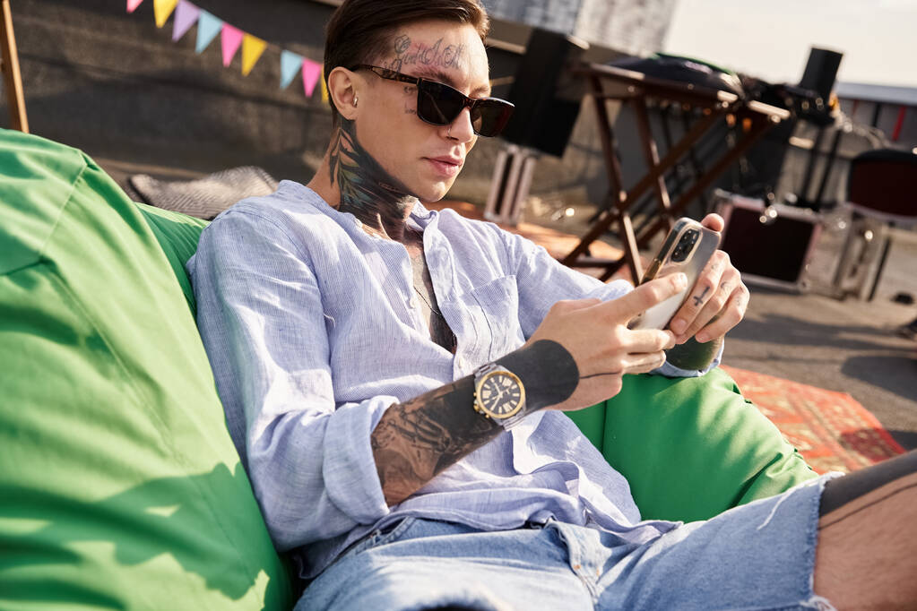 joven guapo con tatuajes y gafas de sol relajante durante la fiesta y mirando el teléfono inteligente - Foto, Imagen