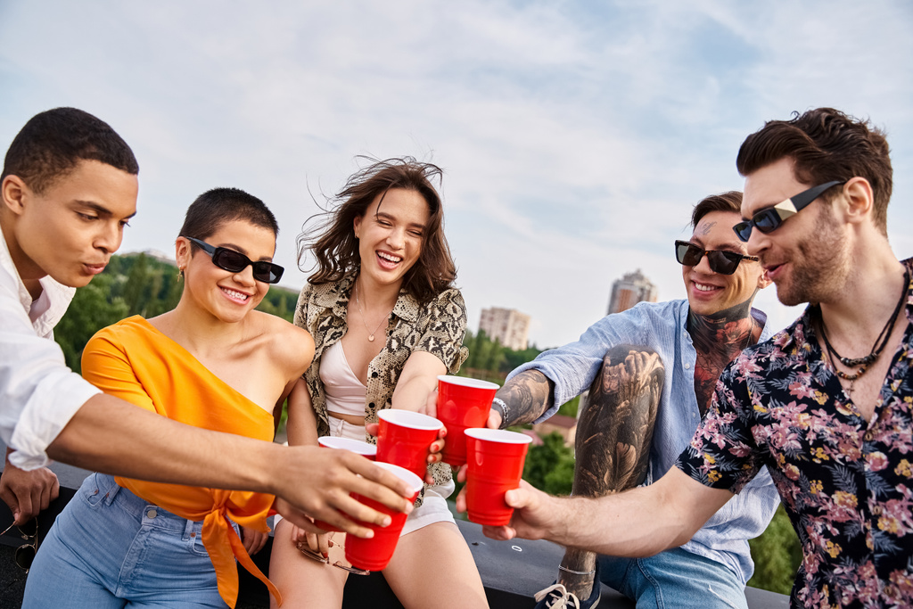 веселые многорасовые друзья в солнечных очках звонят красными чашками с напитками на крыше - Фото, изображение