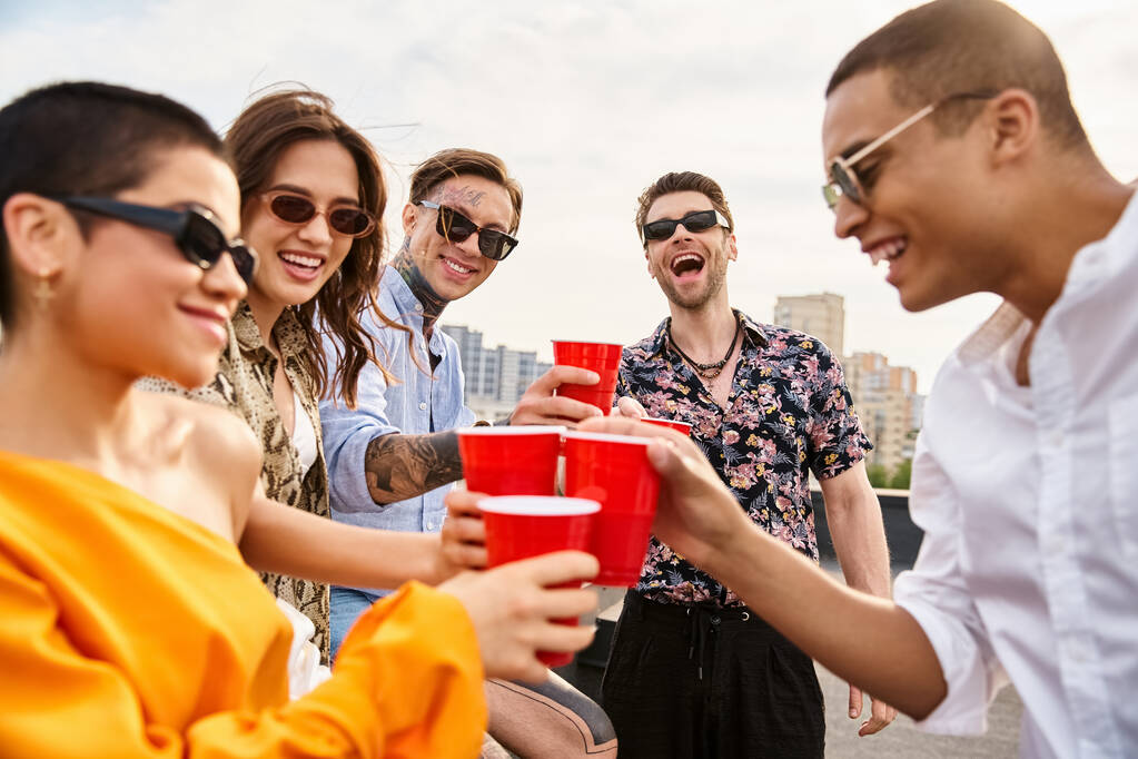 alegres y diversos amigos con gafas de sol divirtiéndose en la fiesta en la azotea sosteniendo copas rojas con bebidas - Foto, imagen