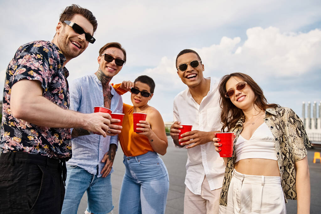 vrolijke interraciale vrienden in levendige kleren met rode bekers met drankjes en glimlachen op de camera - Foto, afbeelding