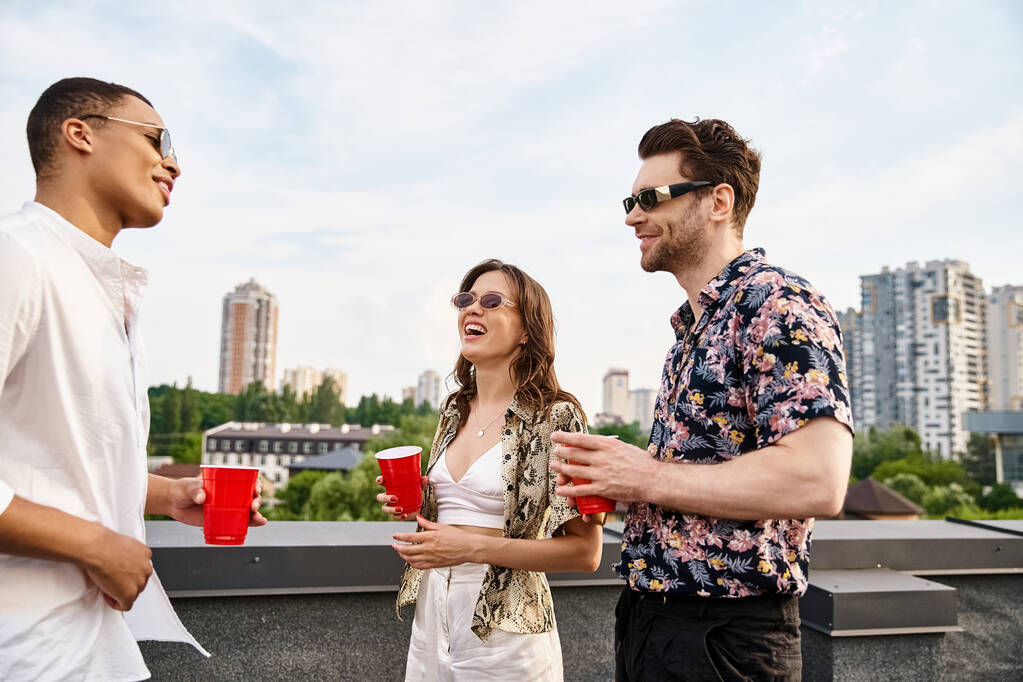 wesołych wielorasowych przyjaciół w stylowych okularach przeciwsłonecznych pijących z czerwonych kubków podczas zabawy na dachu - Zdjęcie, obraz