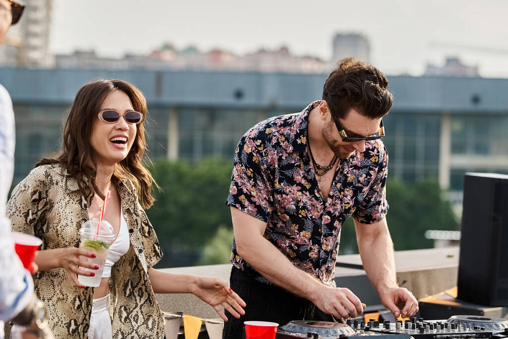 χαρούμενη γυναίκα με κομψά γυαλιά ηλίου και κοκτέιλ στο χέρι ποζάροντας δίπλα στον DJ στο rooftop party - Φωτογραφία, εικόνα