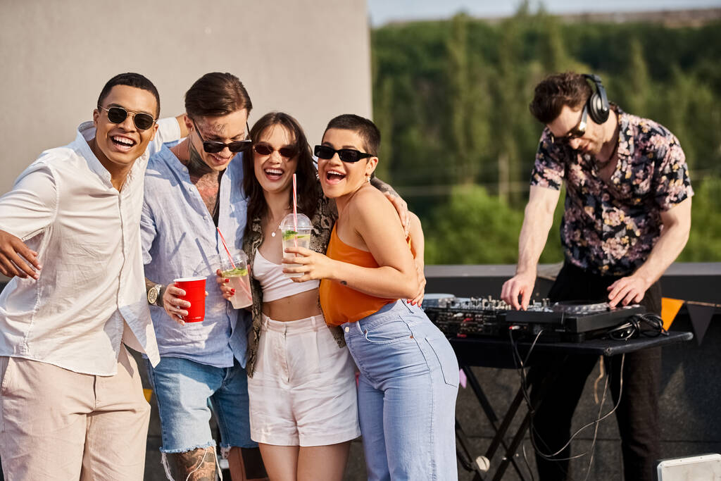 fröhliche interrassische Freunde mit Drinks, die neben DJ auf der Dachparty posieren und in die Kamera lächeln - Foto, Bild