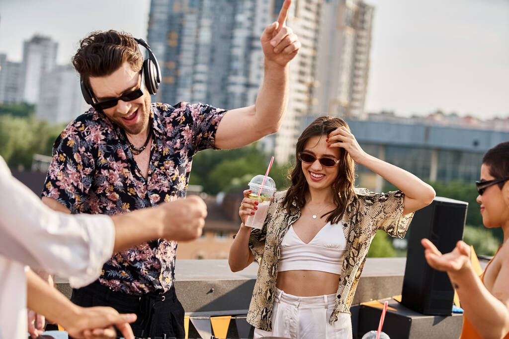 привлекательная веселая женщина с коктейлем в руке, танцующая под DJ сет рядом со своими друзьями на вечеринке на крыше - Фото, изображение