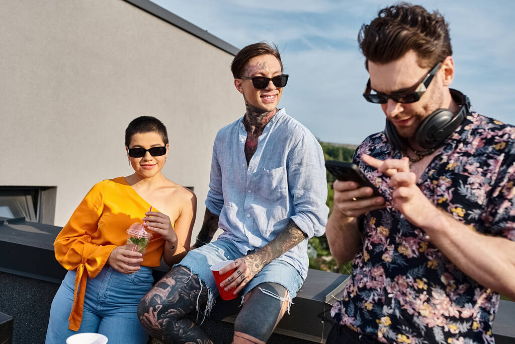 gutaussehender DJ mit Sonnenbrille und Cocktails auf dem Dach neben seinen fröhlichen Freunden auf sein Handy blickend - Foto, Bild