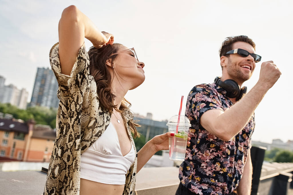 radostná žena s dlouhými vlasy a stylovými slunečními brýlemi držící koktejl vedle DJ na střešní párty - Fotografie, Obrázek