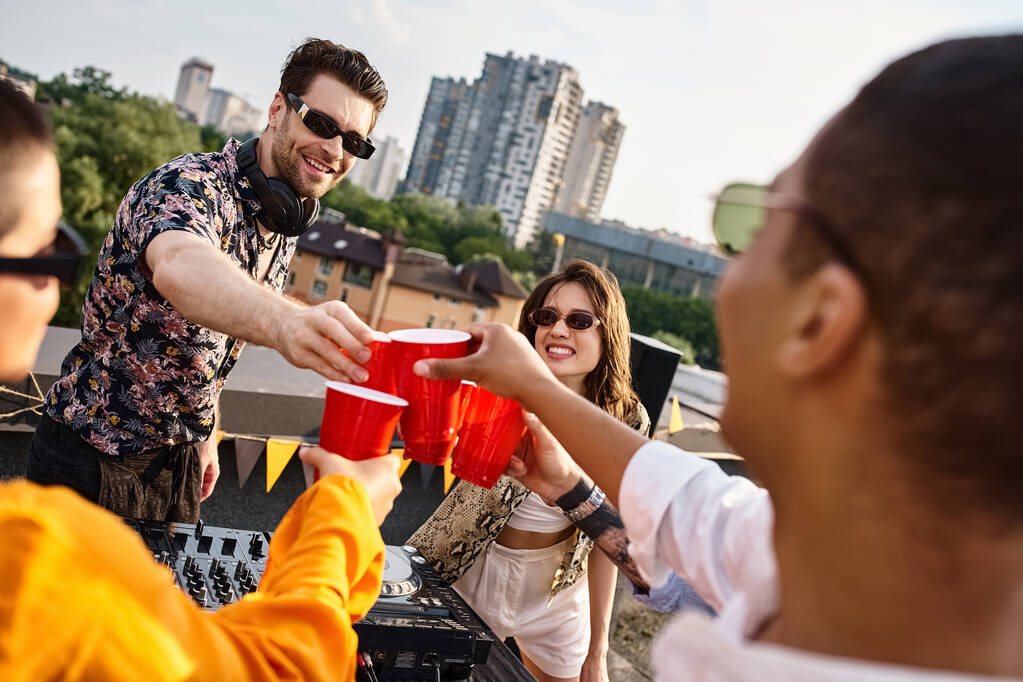 Güneş gözlüklü çok ırklı güzel arkadaşlar çatıdaki partide bardaklarını içkiyle tokuşturuyorlar. - Fotoğraf, Görsel