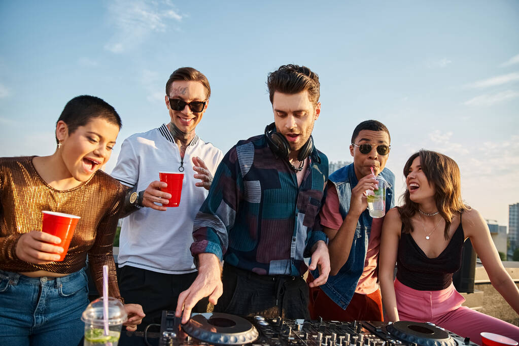interraciale vrolijke mensen in levendige outfits feesten samen naast knappe DJ op het dak partij - Foto, afbeelding