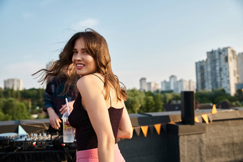 focus op vrolijke aantrekkelijke vrouw kijken naar camera naast haar wazige vrienden op het dak feest - Foto, afbeelding