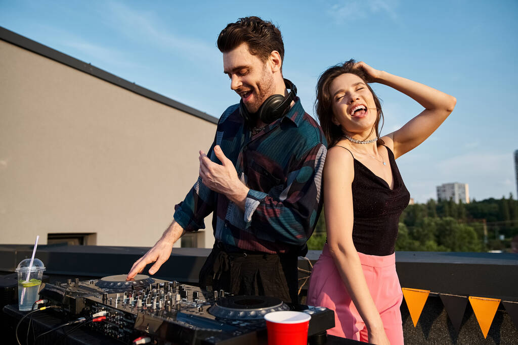 Χαρούμενη ελκυστική γυναίκα με μακριά μαλλιά που διασκεδάζει στο πάρτι στην ταράτσα δίπλα στον όμορφο χαρούμενο DJ - Φωτογραφία, εικόνα