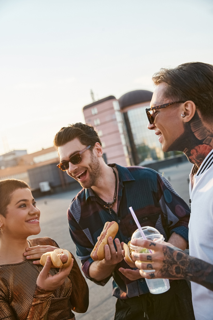 veselé atraktivní přátelé se slunečními brýlemi v městských oblečení těší hot dogy na střešní party - Fotografie, Obrázek