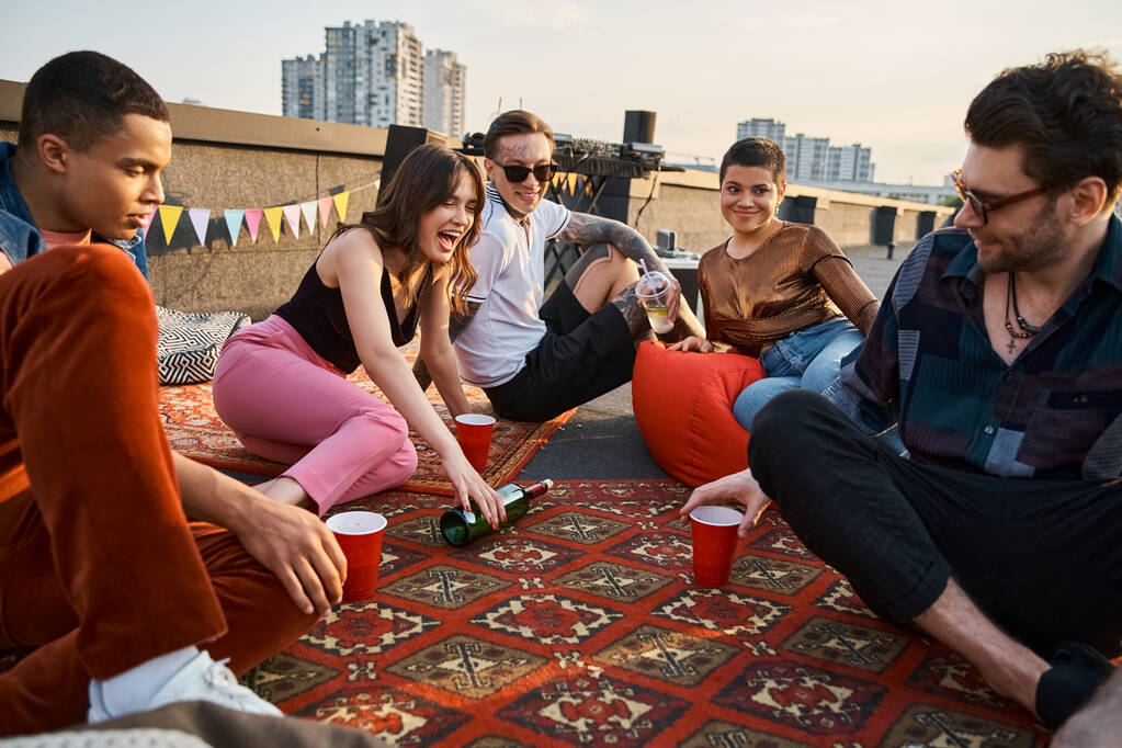 屋上パーティーで一緒にボトルを回して遊ぶ魅力的な陽気な多文化の友人 - 写真・画像