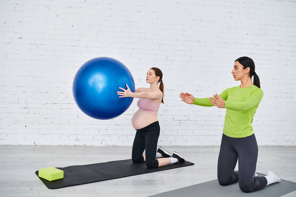 madre embarazada ejercicio con gracia con una pelota durante una clase de fitness prenatal, guiada por su entrenador. - Foto, Imagen