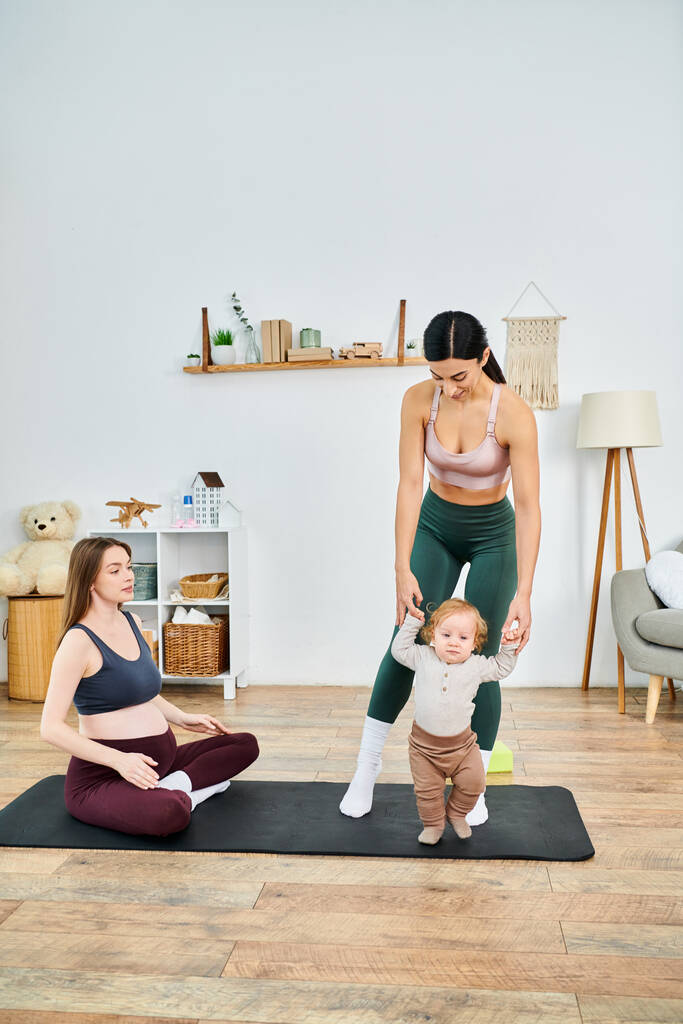 Mladá matka elegantně praktikuje jógu na podložce se svým dítětem, zatímco je vedena svým trenérem na kurzy rodičů. - Fotografie, Obrázek