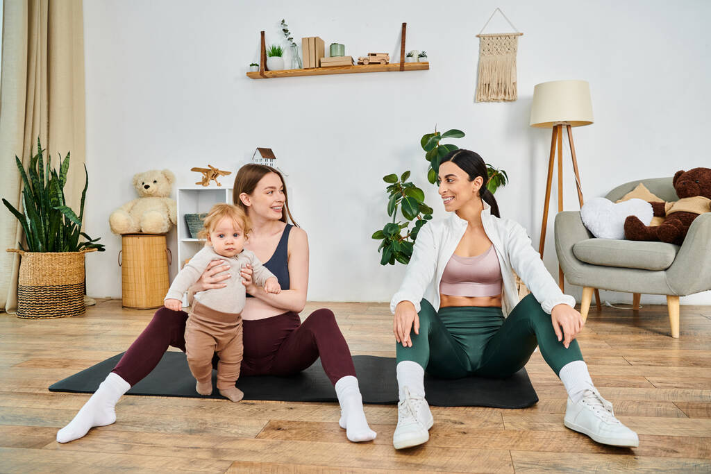 Two women and a baby enjoy a relaxing yoga session together on a colorful mat at home, guided by a supportive coach. - Photo, Image