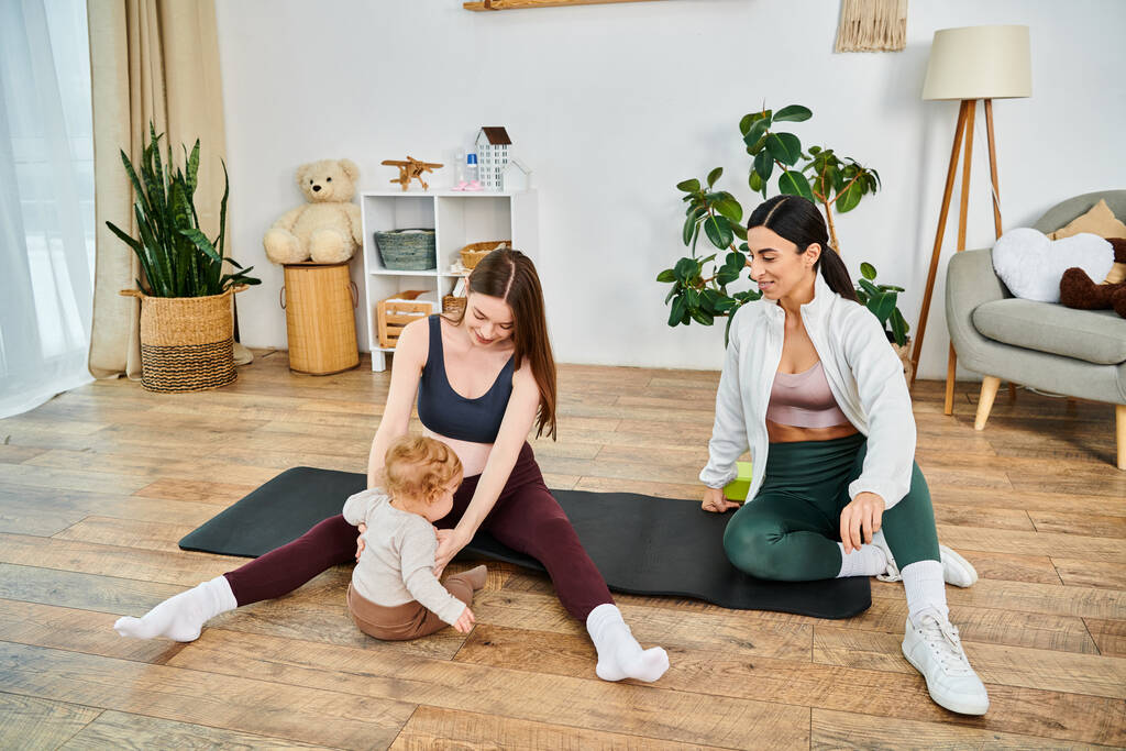 Two women, a serene young mother and her coach, guide a baby on a yoga mat in a peaceful home environment. - Photo, Image