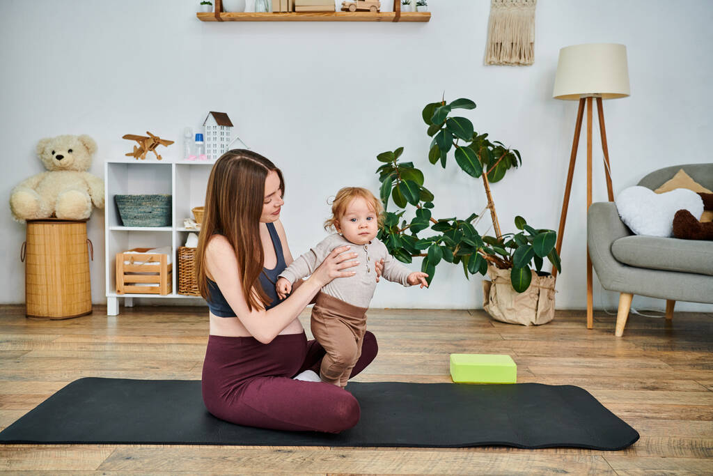 Eine junge schöne Mutter sitzt auf einer Yogamatte und wiegt ihr Baby unter Anleitung ihres Trainers während eines Elternkurses. - Foto, Bild