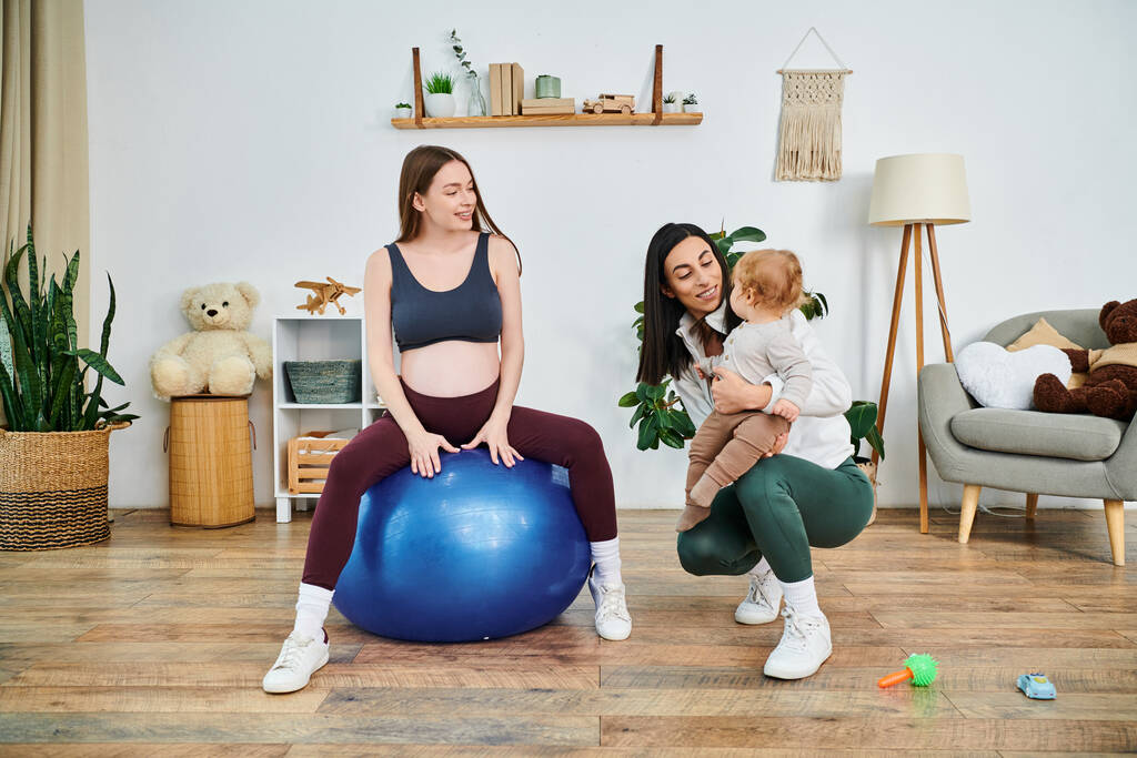 Una joven y hermosa madre se sienta graciosamente en una bola azul, sosteniendo tiernamente a su bebé mientras recibe orientación de su entrenador de crianza.. - Foto, imagen
