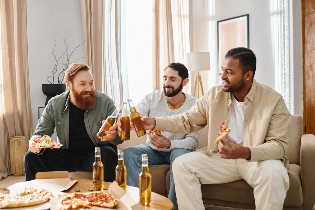 Drei fröhliche, gemischtrassige Männer sitzen auf einer Couch und genießen Pizza und Bier in lockerer Atmosphäre zu Hause. - Foto, Bild