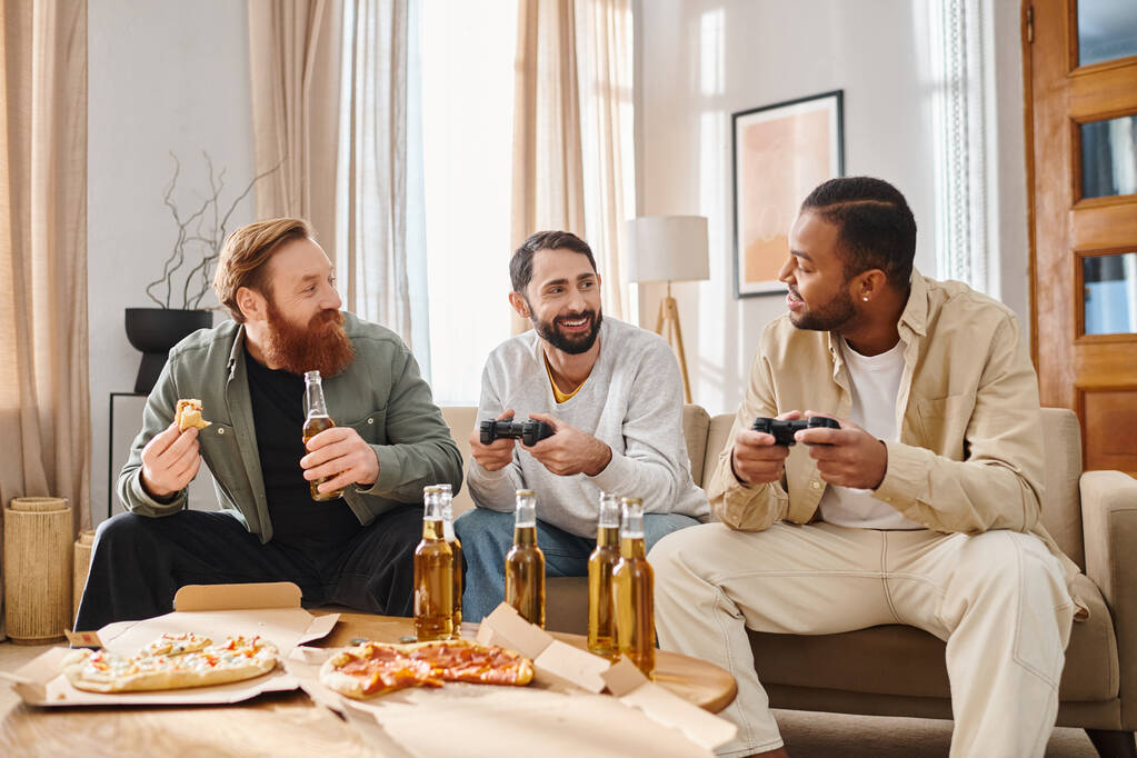 Trzech wesołych, przystojnych mężczyzn o różnym pochodzeniu etnicznym w luźnych ciuchach, łączących się przy grach wideo w domu. - Zdjęcie, obraz