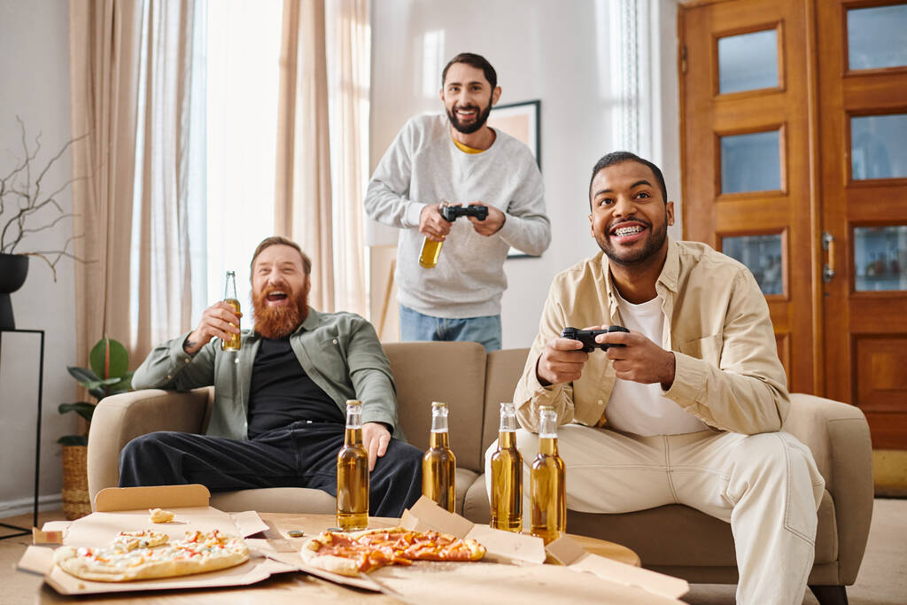Trzech wesołych mężczyzn, różnych ras, śmiejących się i grających w gry wideo na kanapie w luźnych strojach, cieszących się razem zabawą. - Zdjęcie, obraz