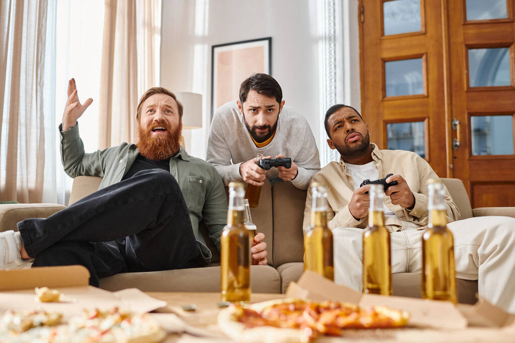 Τρεις διαφυλετικοί όμορφοι άντρες με απλή ενδυμασία κάθονται στον καναπέ γελώντας, τρώγοντας πίτσα και πίνοντας μπύρα.. - Φωτογραφία, εικόνα