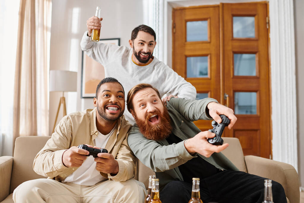 Three handsome men of different ethnicities sit on a couch, smiling and holding remotes, enjoying each others company in a cozy living room. - Photo, Image