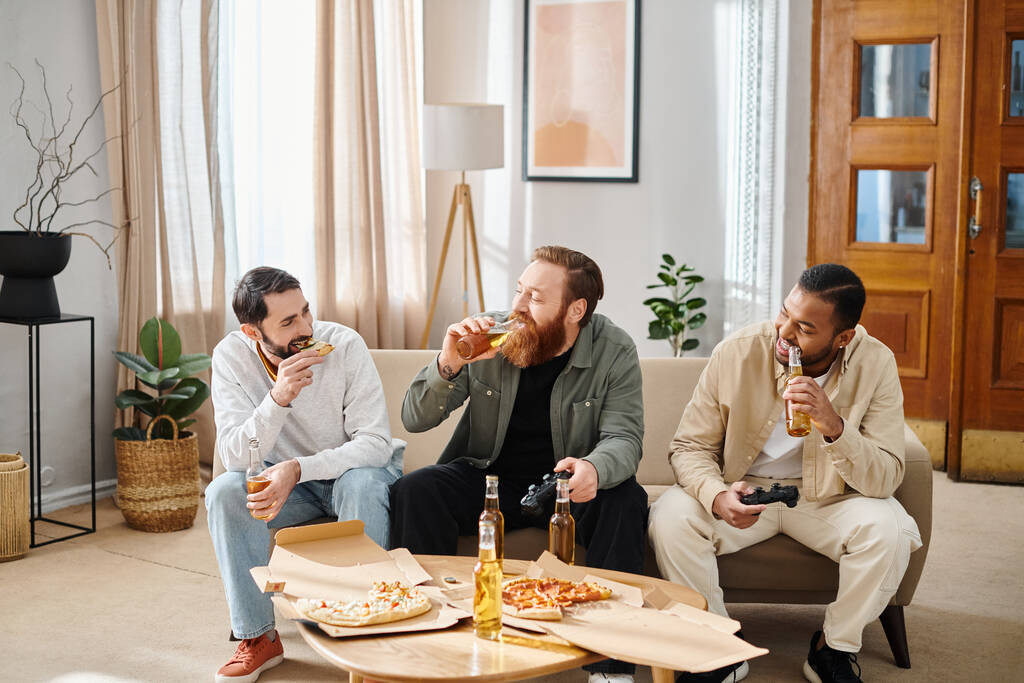 Tres hombres alegres e interraciales en atuendo casual disfrutando de pizza y cerveza en un sofá, expresando amistad y camaradería. - Foto, imagen