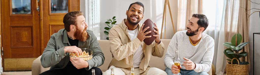 Három változatos férfi ül egy kanapén alkalmi ruhában, boldogan tartva egy focilabdát, élvezve egy vidám és nyugodt pillanatot együtt.. - Fotó, kép