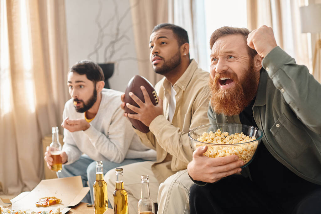 Tři veselí, různorodí muži v neformálním oblečení sledující fotbal a svačící na popcorn v útulném prostředí. - Fotografie, Obrázek