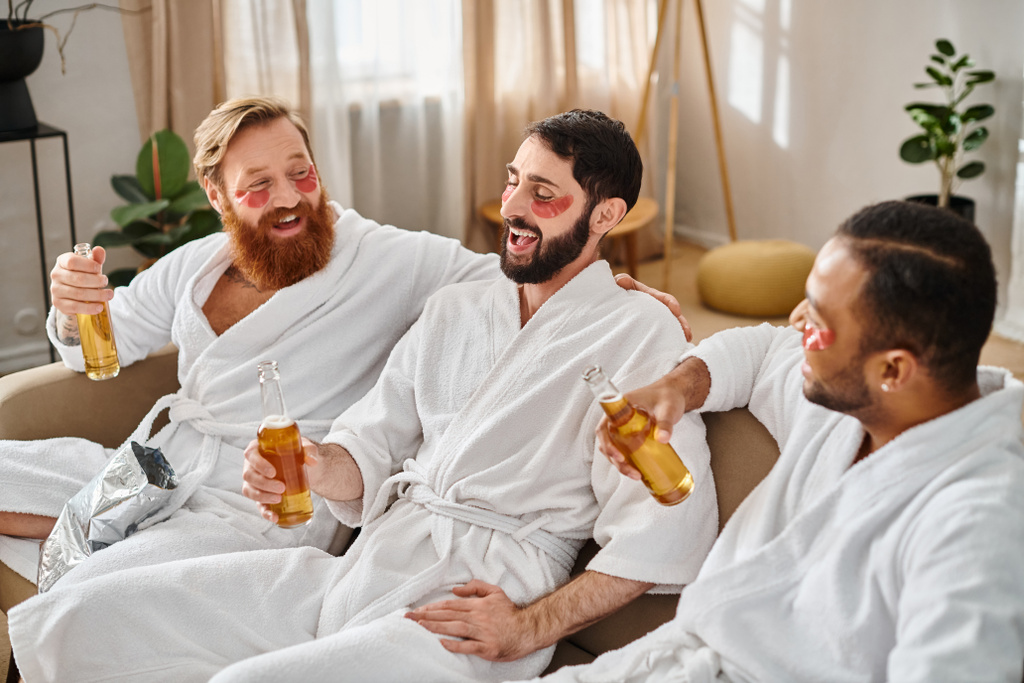 Drei unterschiedliche, gut gelaunte Männer in Bademänteln genießen eine tolle Zeit zusammen, während sie auf einer Couch sitzen. - Foto, Bild