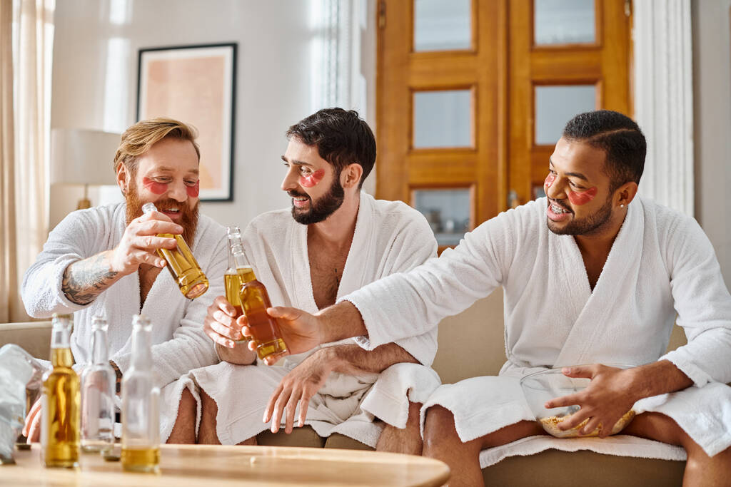Diversi uomini in accappatoi si siedono su un divano, ridendo e gustando bevande dalle bottiglie in un ambiente accogliente. - Foto, immagini