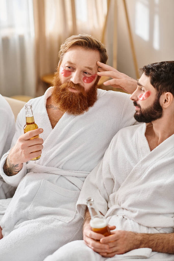 Δύο άνδρες με μπουρνούζια, χαμογελαστοί, κάθονται σε έναν καναπέ κρατώντας μπουκάλια μπύρας, απολαμβάνοντας ο ένας τον άλλο παρέα και φιλία. - Φωτογραφία, εικόνα