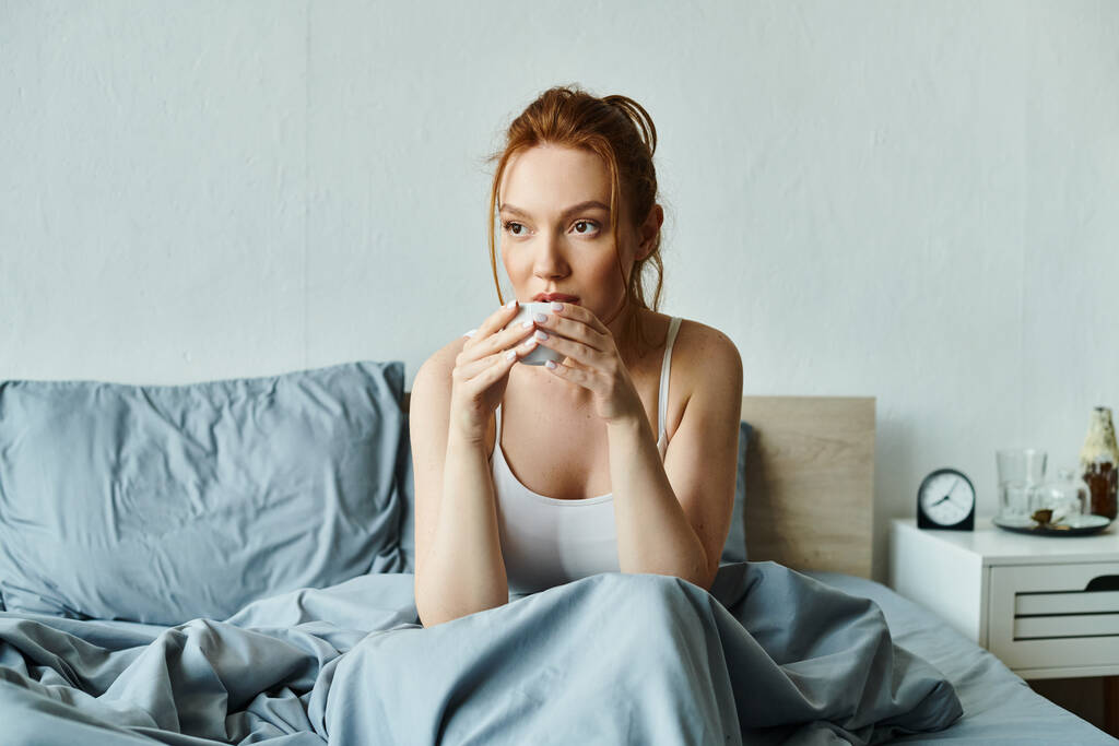 Μια γυναίκα με κομψή ενδυμασία κάθεται σε ένα κρεβάτι με μια μπλε κουβέρτα, εκφράζοντας ικανοποίηση. - Φωτογραφία, εικόνα