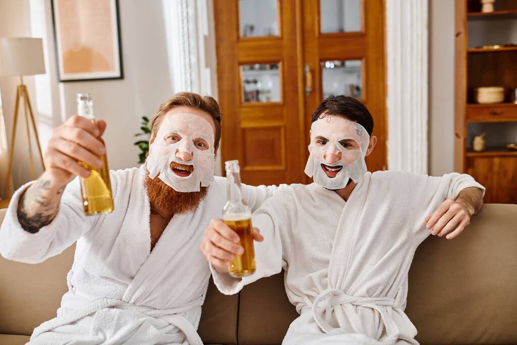 Two men in white robes share a fun moment, holding beer and wearing facial masks for a relaxing and enjoyable time together. - Photo, Image