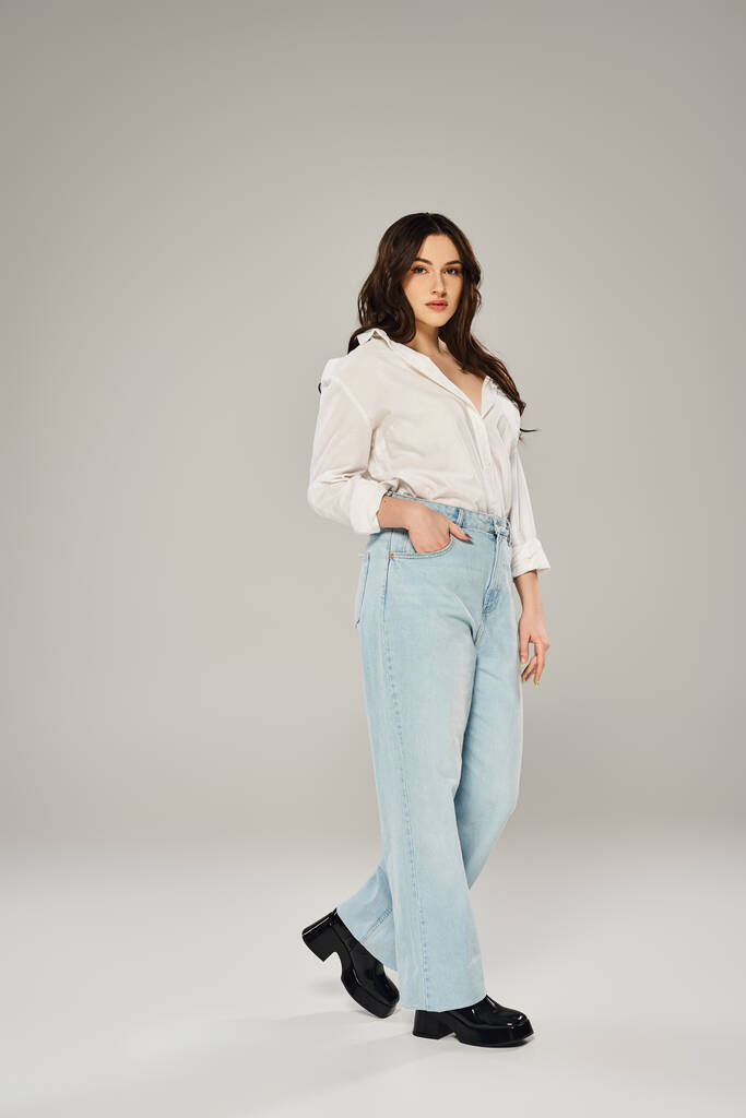 Elegante mulher plus size posando confiantemente em camisa branca e jeans azuis em um pano de fundo cinza. - Foto, Imagem
