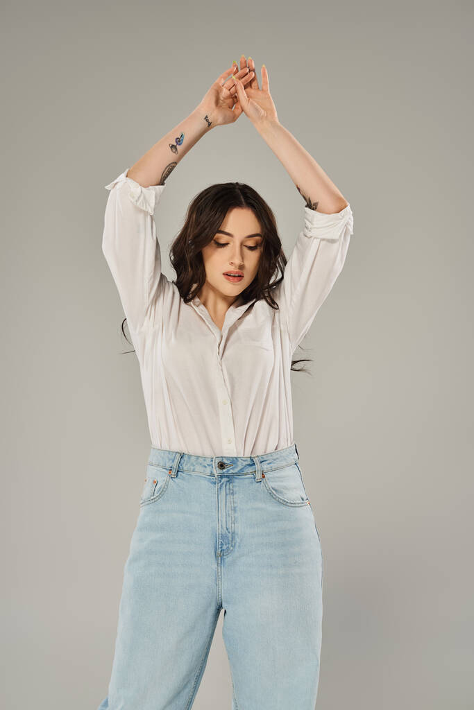 Eine atemberaubende Plus-Size-Frau posiert selbstbewusst in einem stylischen weißen Hemd und blauen Jeans vor grauem Hintergrund. - Foto, Bild