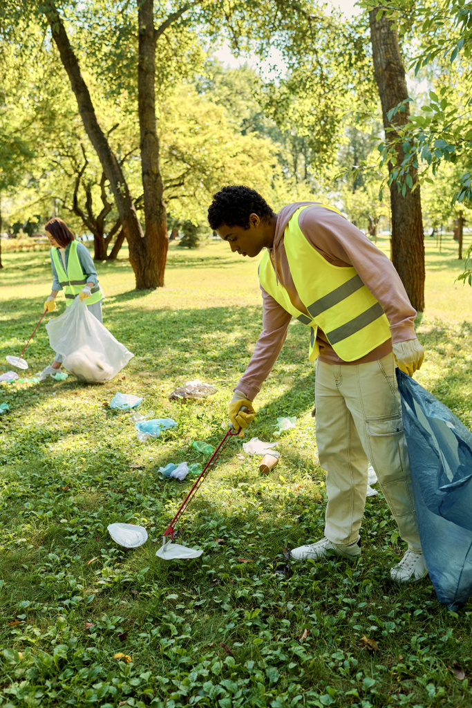 African american man in a yellow safety vest cleans the grass in a park, working alongside a his wife, socially active couple in gloves. - Photo, Image