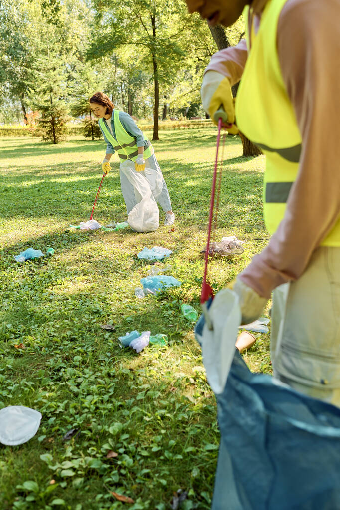Een sociaal actief, divers liefdevol koppel in veiligheidsvesten en handschoenen reinigt een park terwijl een groep op een weelderig groen veld staat. - Foto, afbeelding