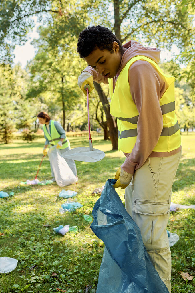 Zróżnicowana para w żółtej kamizelce trzyma niebieską torbę, podczas gdy uczestniczy w sprzątaniu parku z aktywną społecznie, zróżnicowaną kochającą się parą. - Zdjęcie, obraz